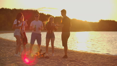 The-students-are-dancing-at-sunset-on-the-sand-beach-in-shorts-and-t-shirts-around-bonfire-with-beer.-They-are-drinking-beer-and-enjoying-the-warm-summer-evening-near-the-river.